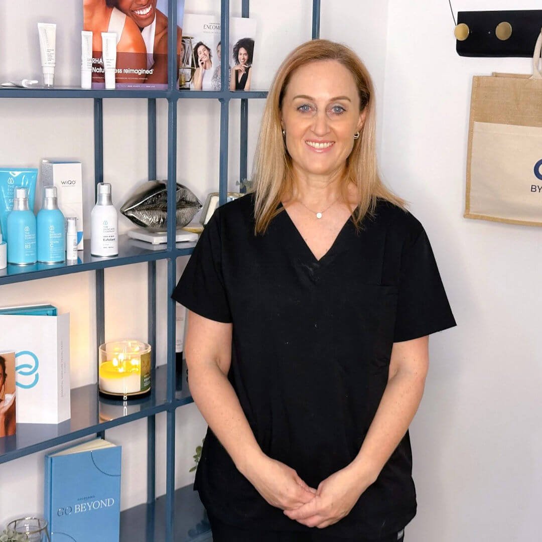 RN Jolieta is wearing black scrubs, standing in a skincare or beauty clinic with shelves of skincare products and magazines behind her.