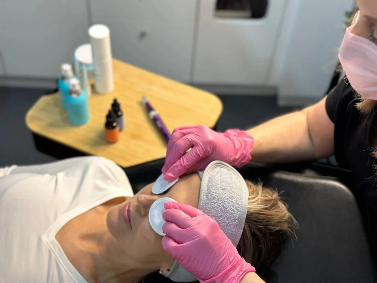A client receives a facial treatment while lying down, with RN Jolieta wearing pink gloves and applying a product to their face in a clinical setting with skincare bottles on a table.