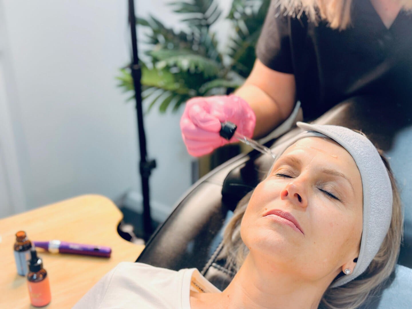 A client participating in a cosmetic treatment procedure, lying on a bed with her eyes closed, receiving a PRX Plus treatment from RN Jolieta wearing pink gloves.