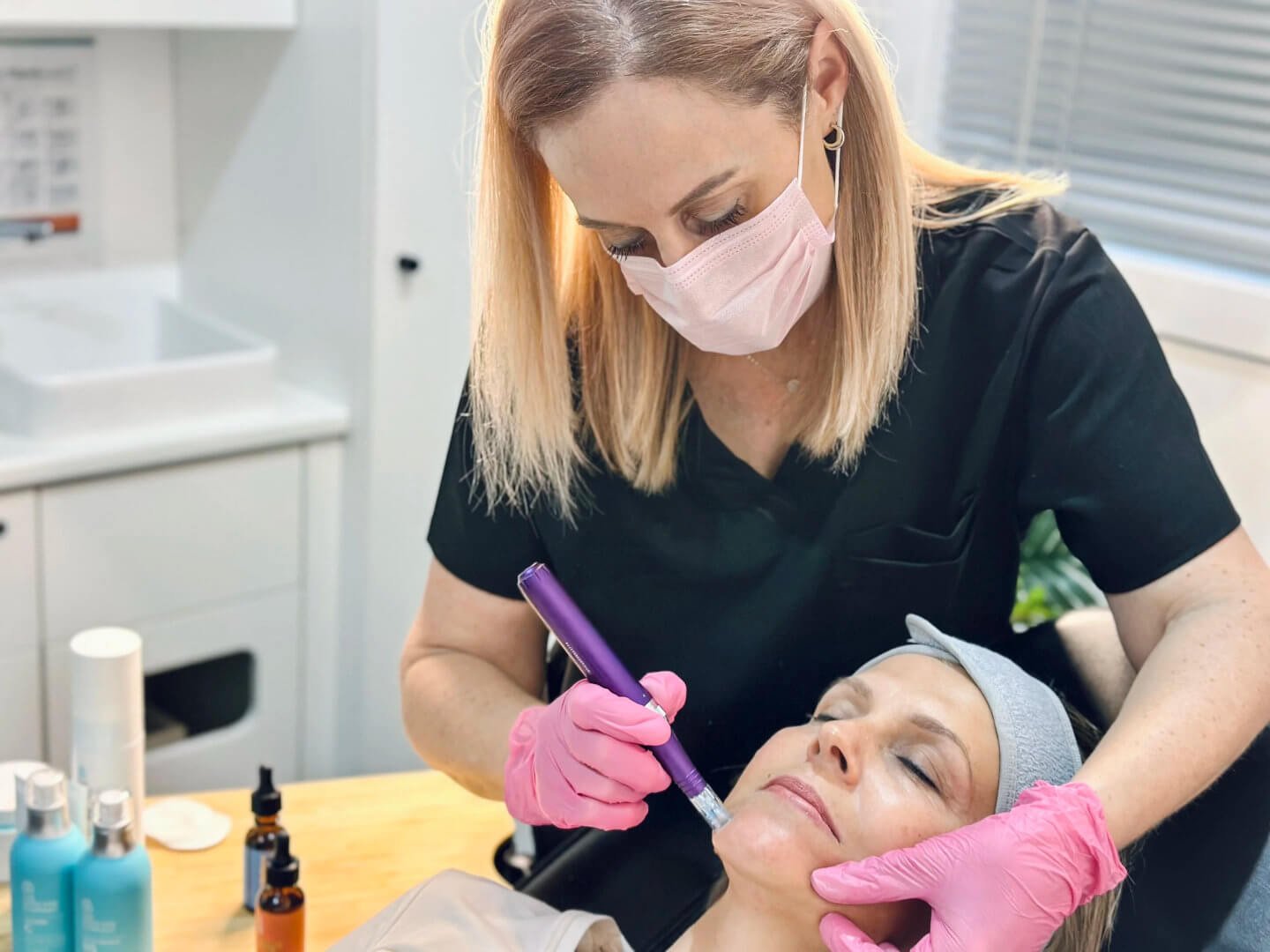 RN Jolieta, wearing a pink face mask and pink gloves, performs a cosmetic procedure on a client lying on the treatment bed in the clinic.