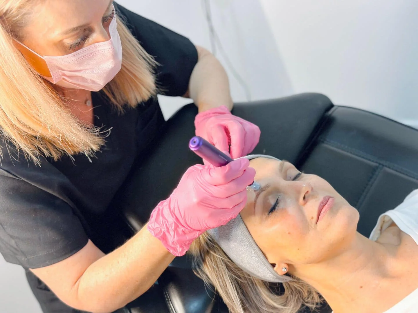 A client is lying down with her eyes closed while RN Jolieta, wearing pink gloves and a face mask, performs microneedling.