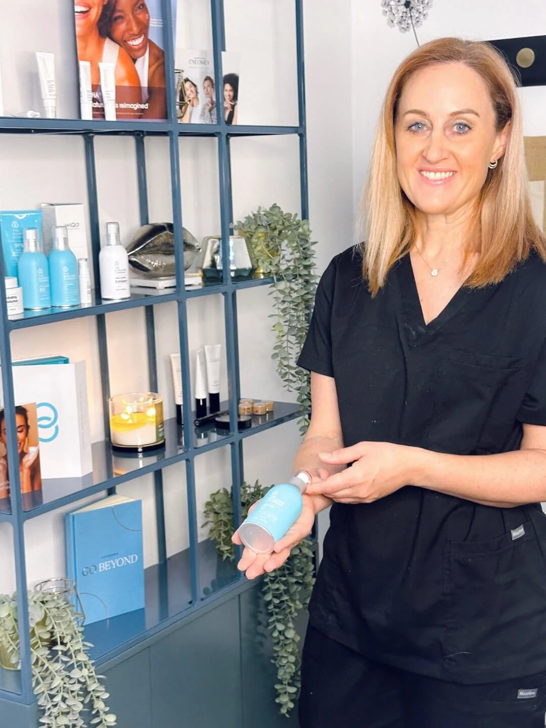 RN Jolieta in black scrubs, smiling and holding a blue skincare bottle inside a skincare clinic or store, with shelves of skincare products, plants, and promotional posters in the background.