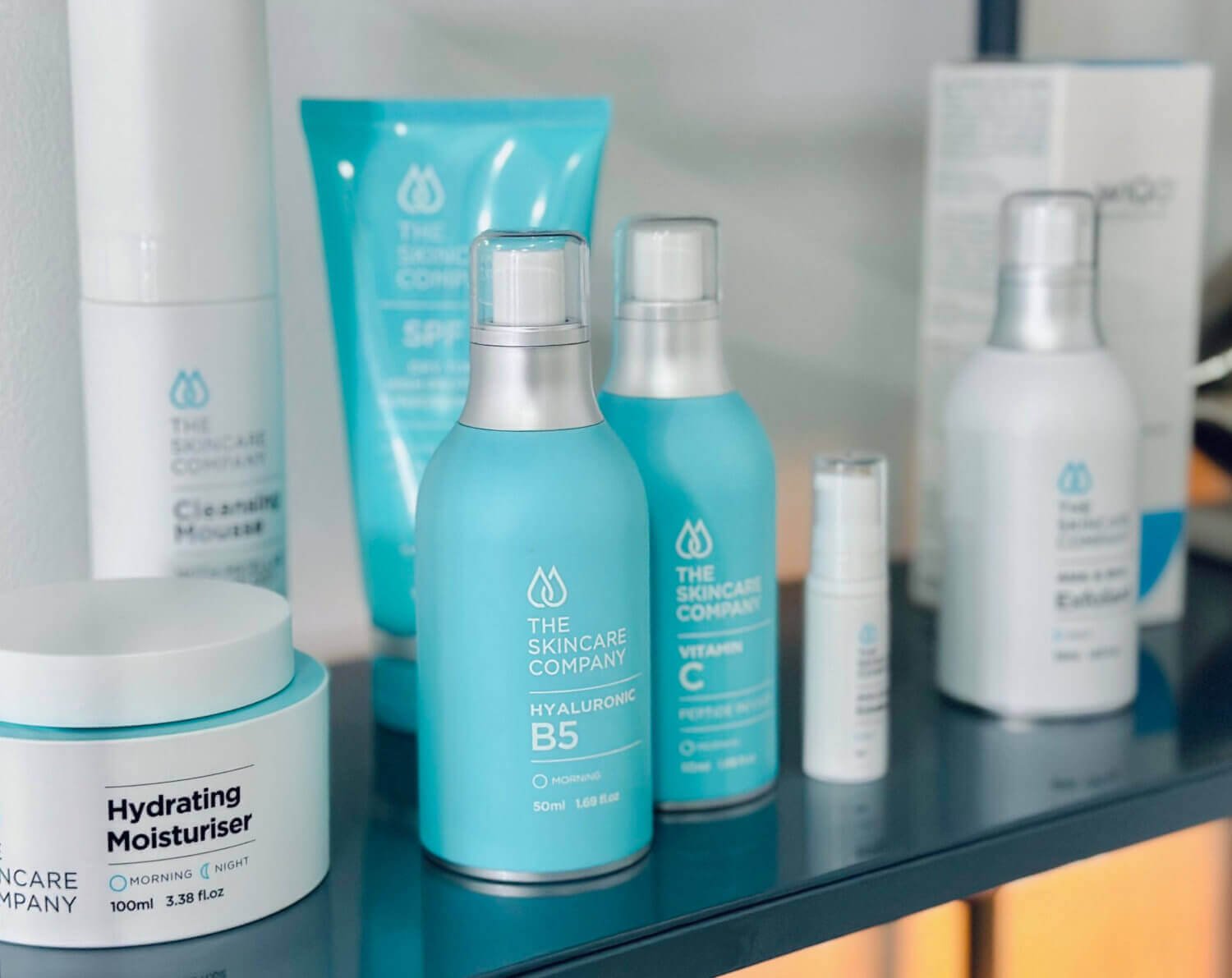 Skincare products on a black shelf, including bottles labeled 'The Skincare Company,' moisturer and hyaluronic B5, with a tube of cream in the background