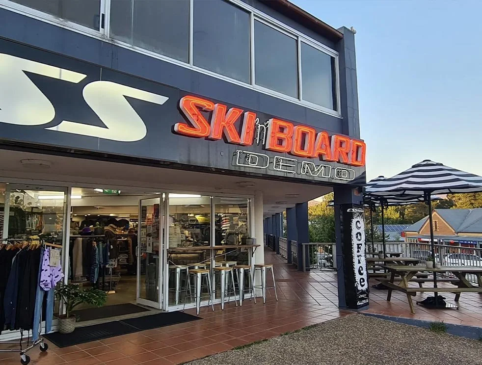 Exterior of a building with a large neon sign that reads 'SK8 BOARD' and a smaller sign that says 'DEMO.' The entrance has glass doors and windows, with clothing racks inside. Outside, there are picnic tables with an umbrella, and a sign that reads 'COFFEE' next to the entrance. The sky is clear and it appears to be early evening.
