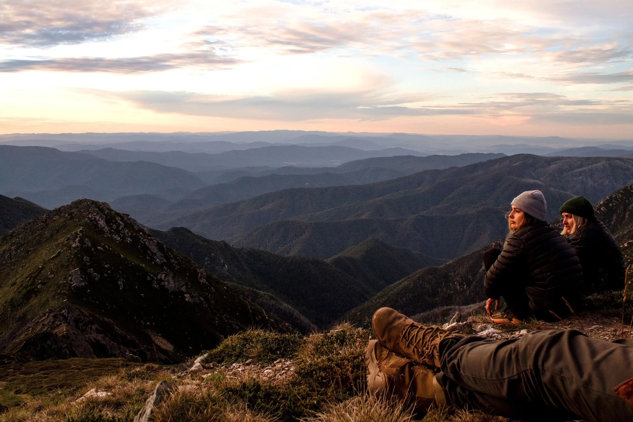 Two women sitting on a mountain ridge watching a sunset over layered mountain ranges, with hiking boots and gear visible in the foreground.