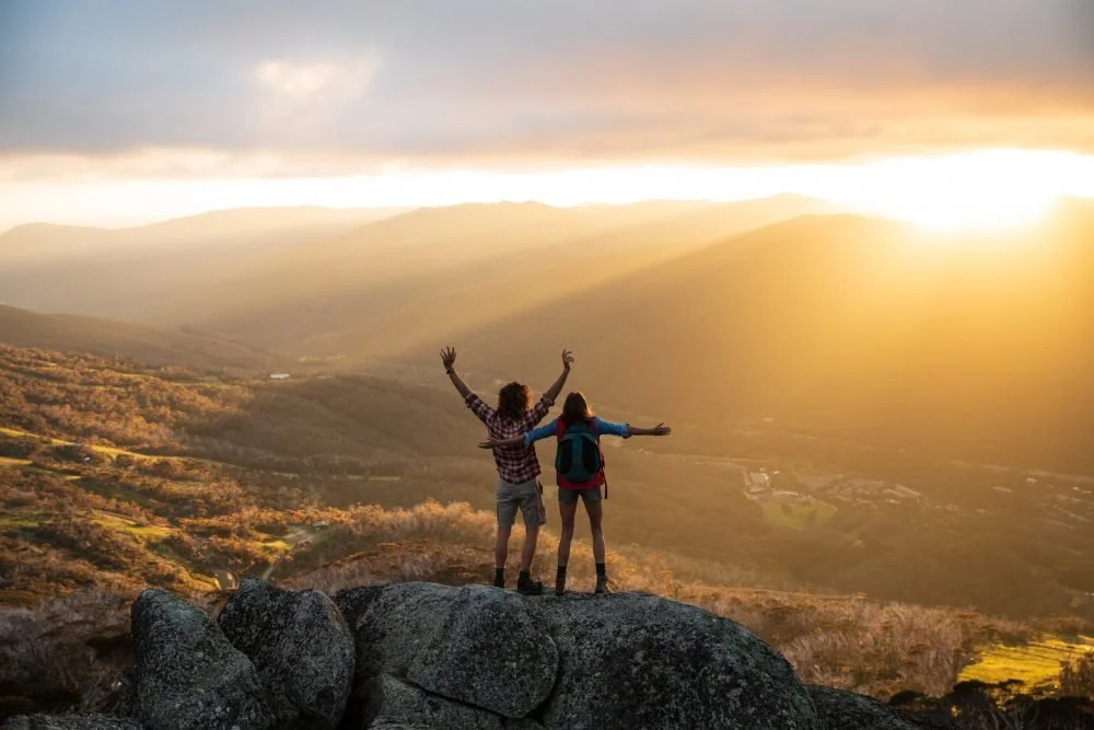 Two friends standing on a large rock at sunset, overlooking a valley with mountains in the distance, with their arms raised.