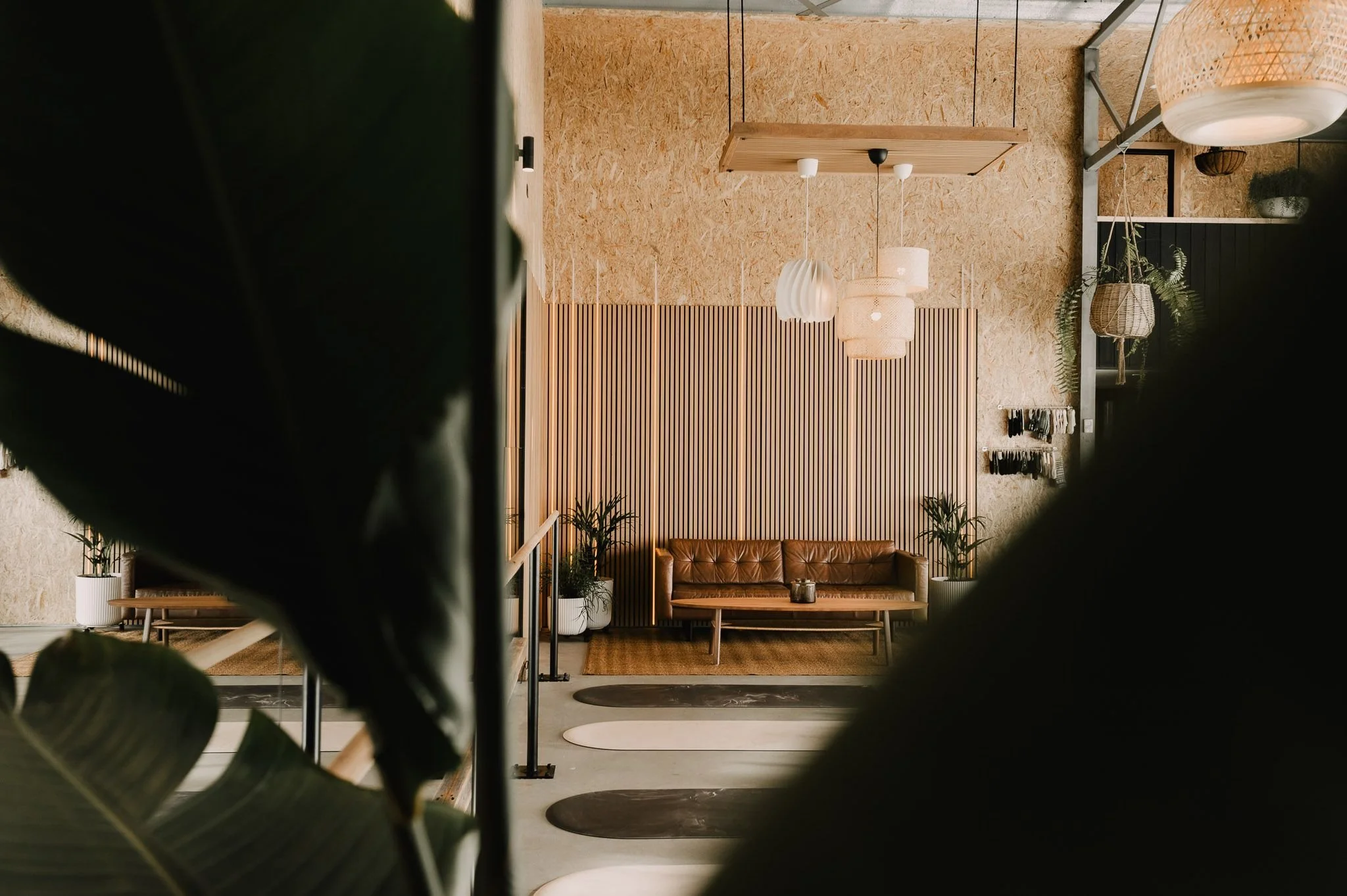 Cozy lobby with a leather sofa, potted plants, hanging pendant lights, and a textured wooden wall, seen through large dark leaves.