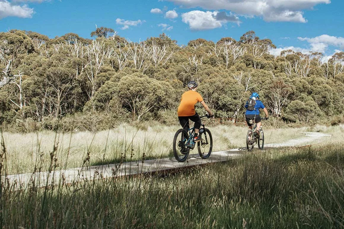 Two people riding bicycles on a paved trail through a grassy, wooded area during daytime with a blue sky and scattered clouds.