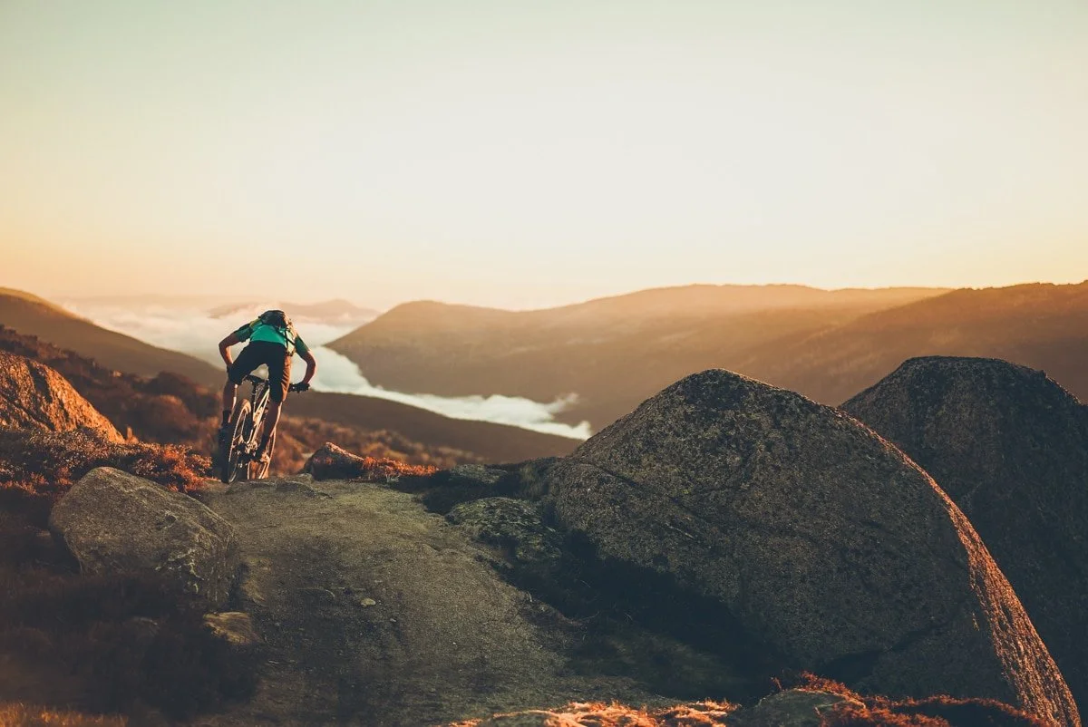 A mountain biker riding on a trail through rocky terrain with mountains and clouds in the background during sunset or sunrise.
