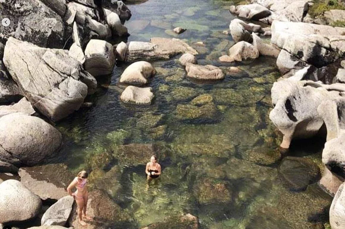 Two people, one standing and one sitting, in a rocky, shallow mountain stream surrounded by large rocks.