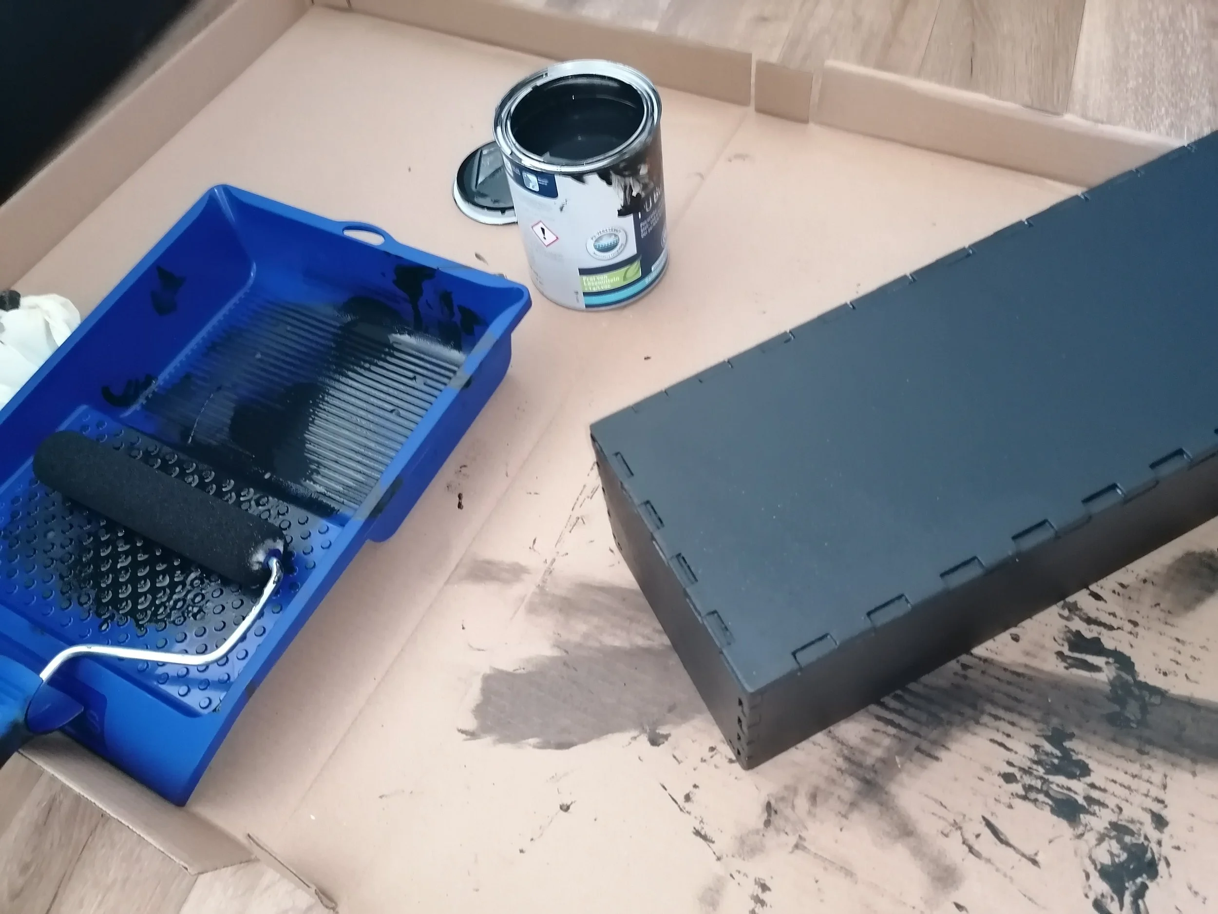 A paint roller and tray, an open can of black paint, and a black toolbox placed on a cardboard surface, indicating a painting project in progress.