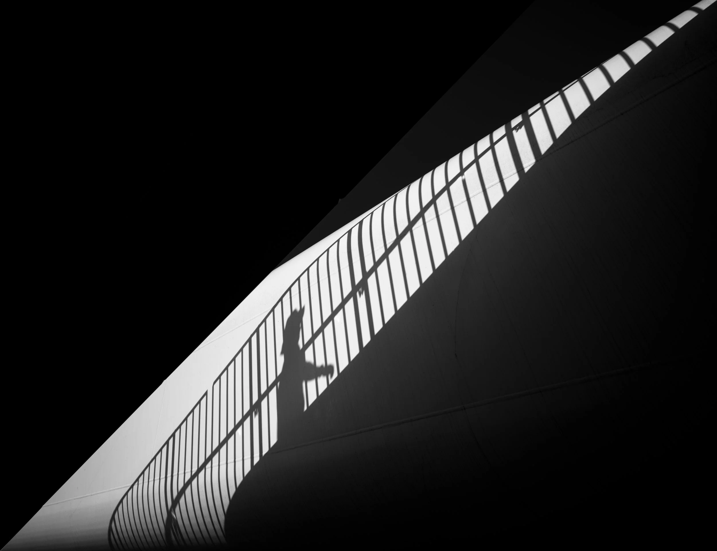 A black and white photo of a person's shadow walking on a curved bridge with a railing, cast by sunlight.