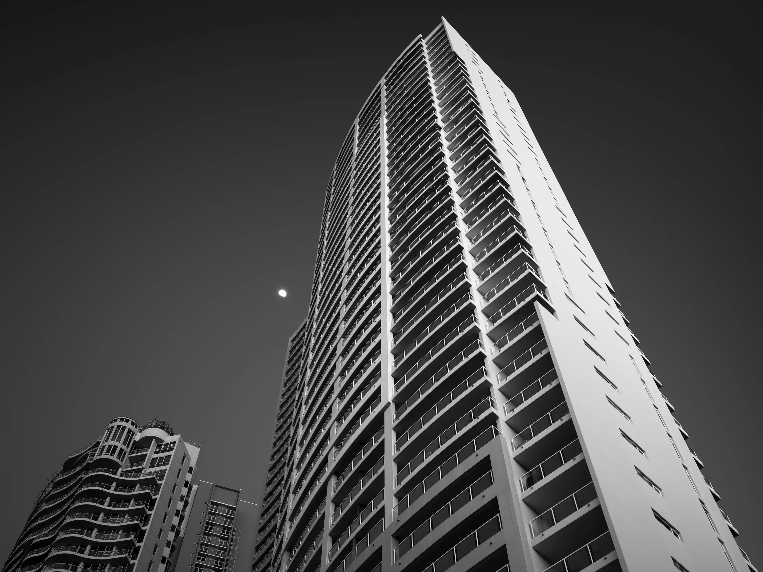 Black and white photo of tall modern skyscraper with numerous balconies, viewed from a low angle against a dark sky with visible moon.