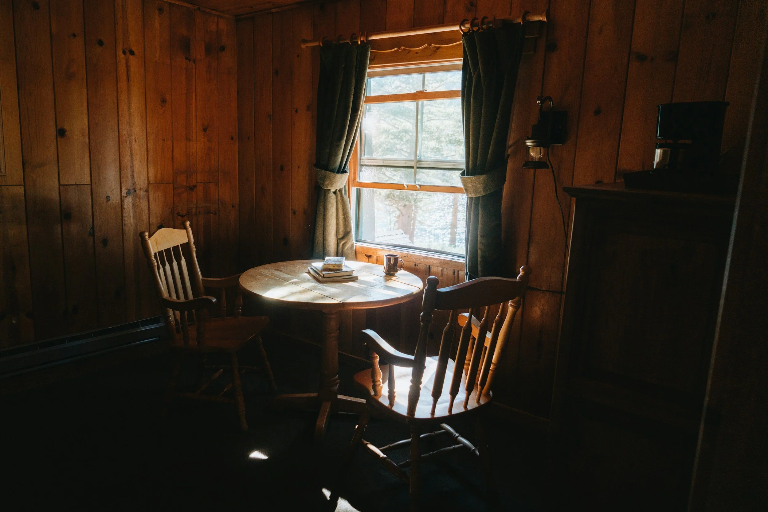 A cozy wooden room with sunlight streaming through a window, a round wooden table with books and a mug, and two wooden chairs.