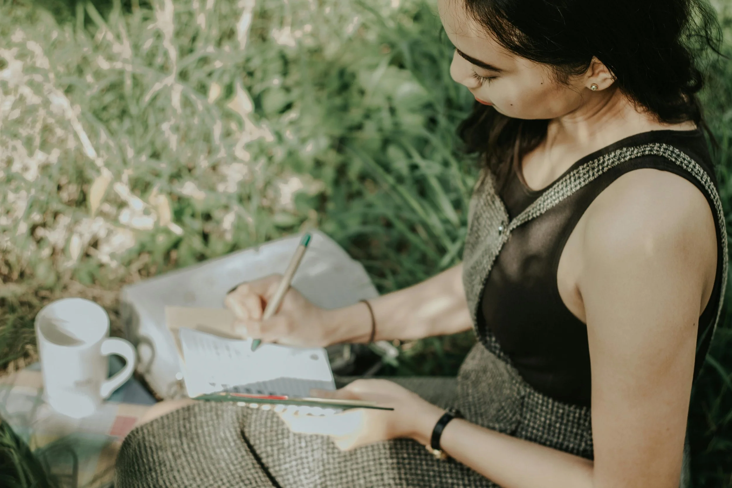 A woman sitting outdoors on lush green grass, writing in a notebook with a pen, surrounded by a white mug and other items.