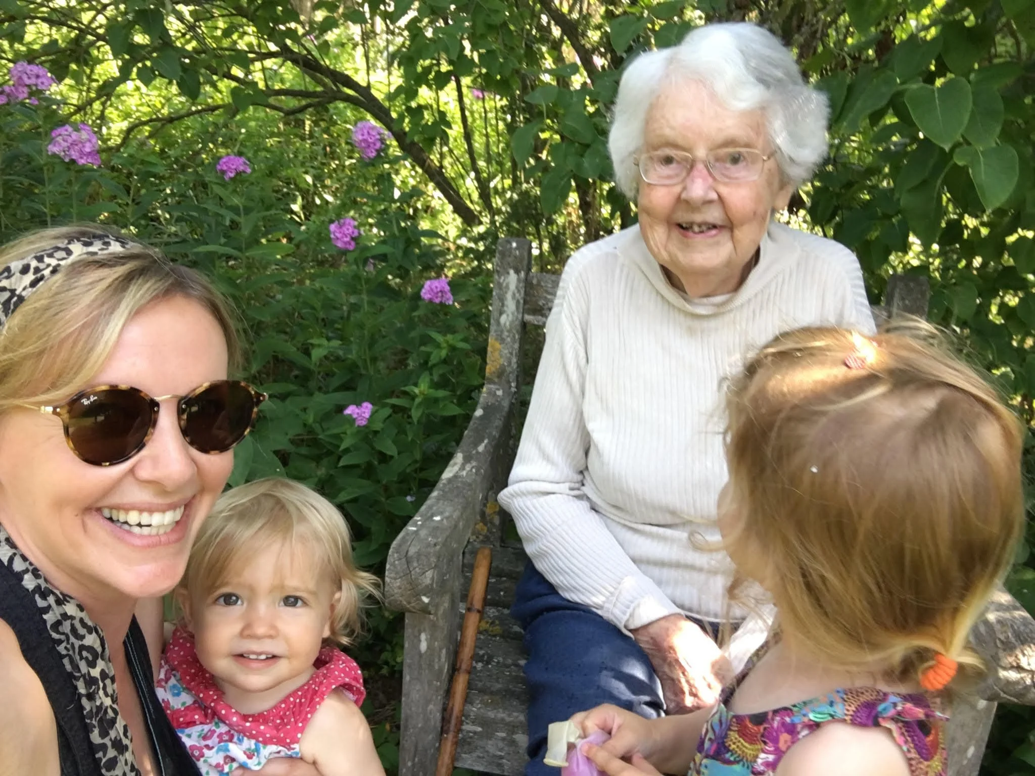 Four people, including a smiling elderly woman sitting on a garden bench and two young children with their mother, surrounded by green foliage and purple flowers.