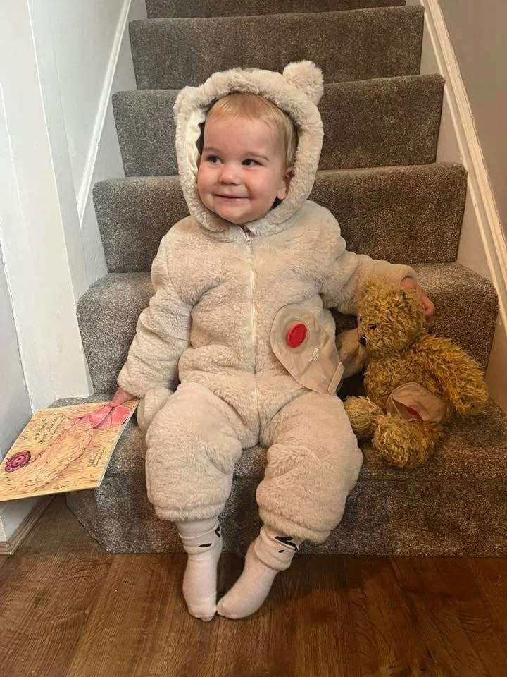 A young child dressed in a fluffy, beige bear costume sitting on a staircase, holding a teddy bear, with a book on the floor beside them, smiling.