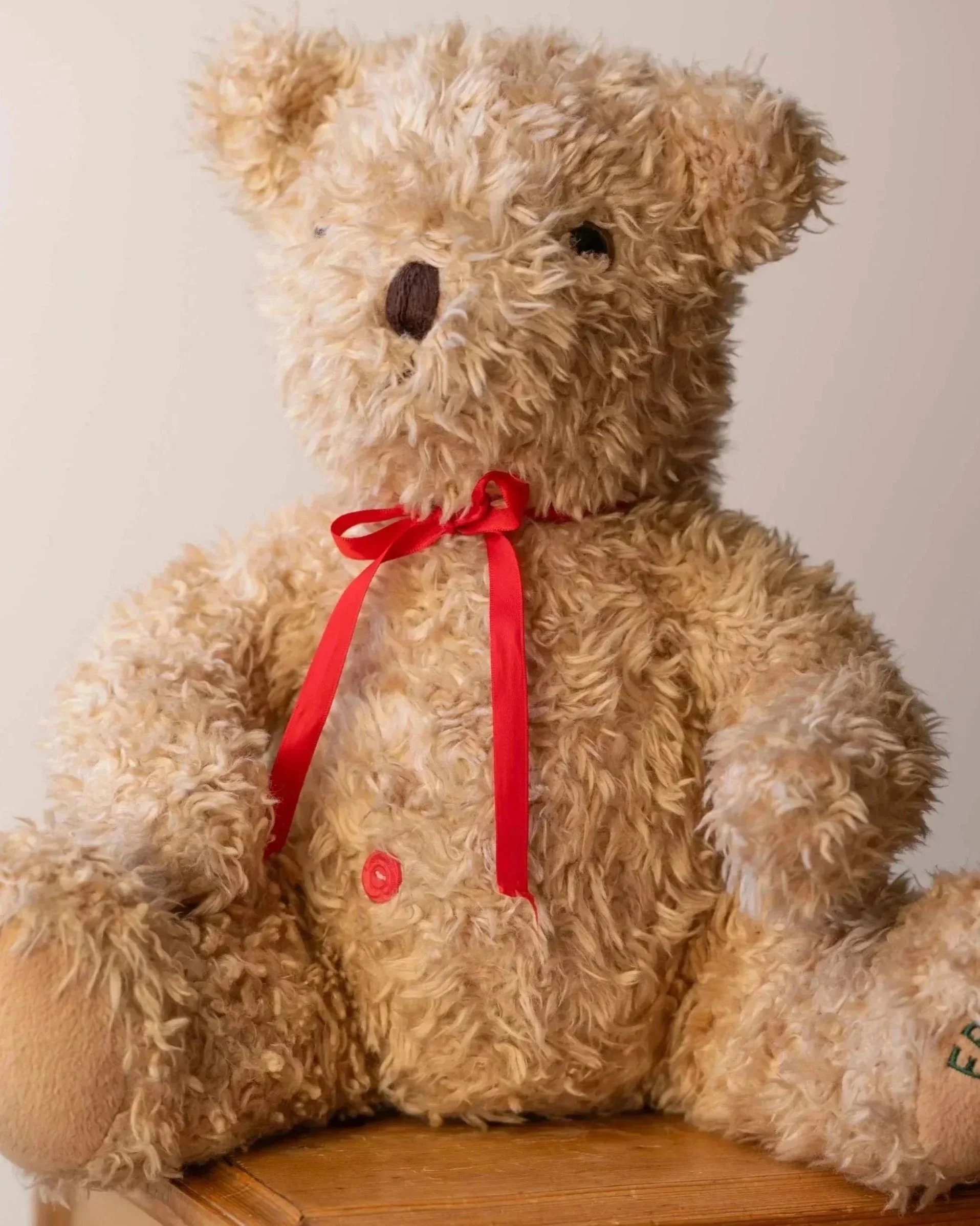 A fluffy beige teddy bear with a red ribbon around its neck, sitting on a wooden surface against a plain background.
