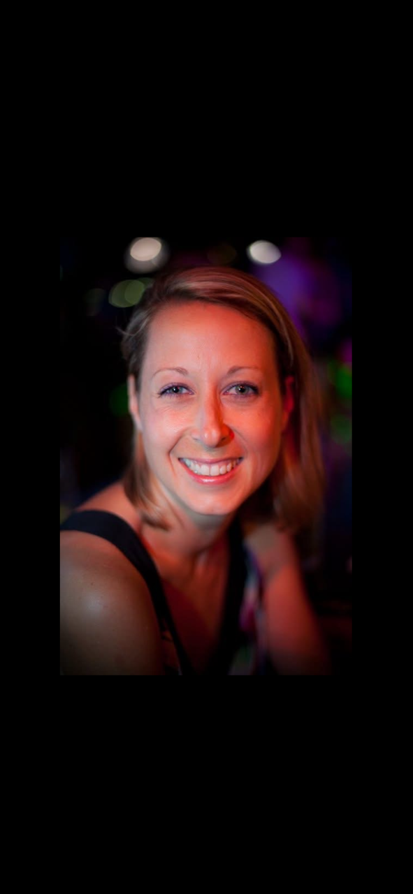 A woman smiling at the camera in a dimly lit environment with colorful blurred lights in the background.