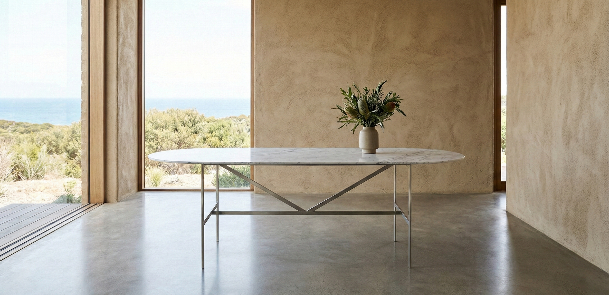 Minimalist interior with a round marble-top table and a vase with flowers, large glass sliding door with a view of greenery and the ocean in the background.