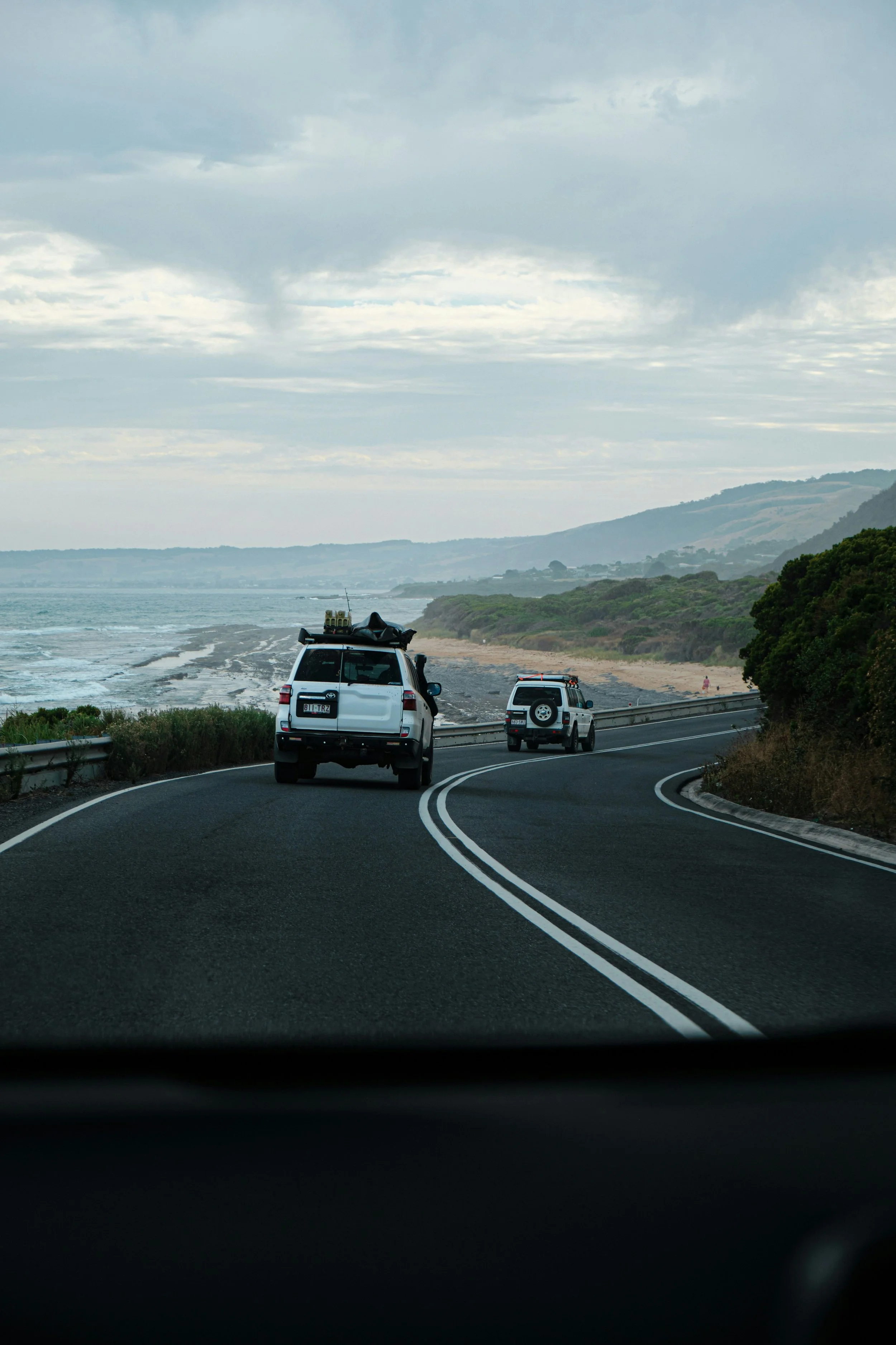 Vehicles driving along a scenic coastal road with ocean on the left and hills on the right under cloudy sky.