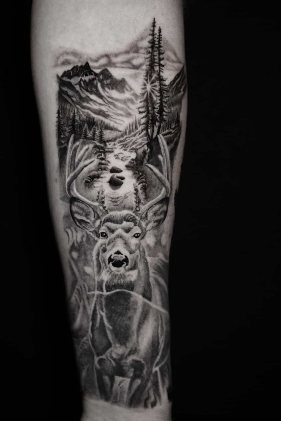 Black and gray tattoo on arm depicting a nature scene with mountains, trees, a river, and a deer with large antlers.