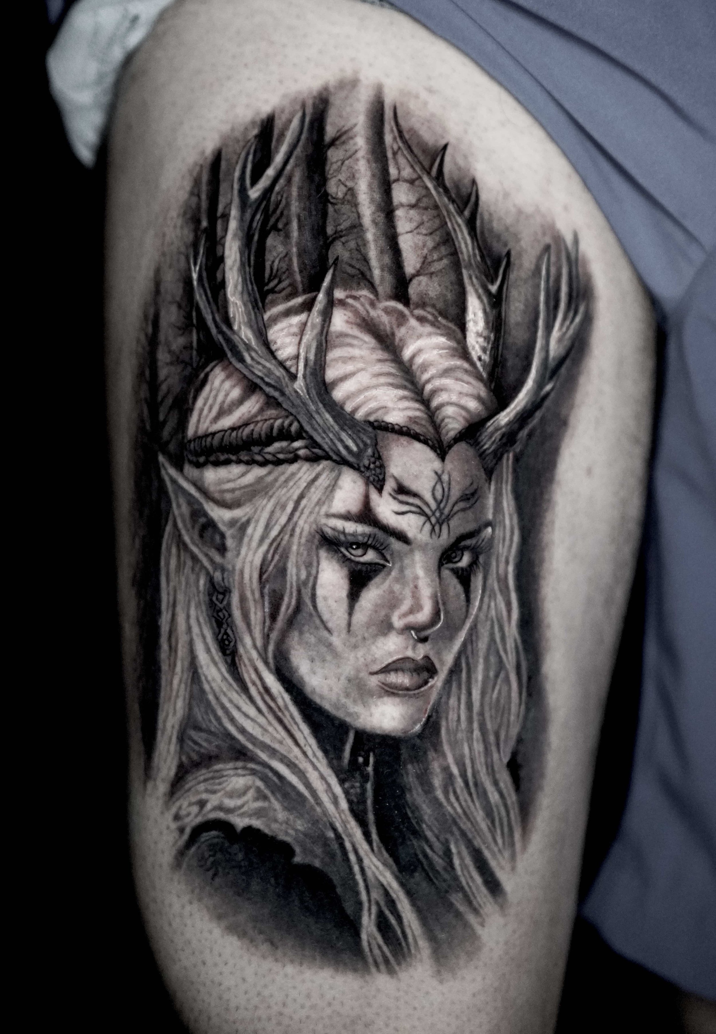 Black and gray tattoo of a fantasy woman with deer antlers, long hair, and face paint, on upper arm.