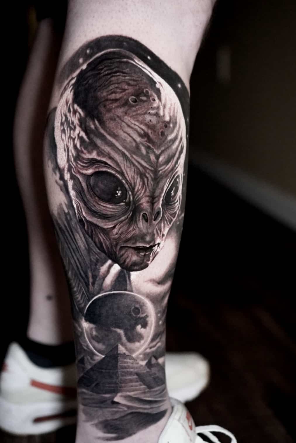Tattoo of an alien face with large eyes and textured skin, moon and pyramid in the background, on a person's leg.