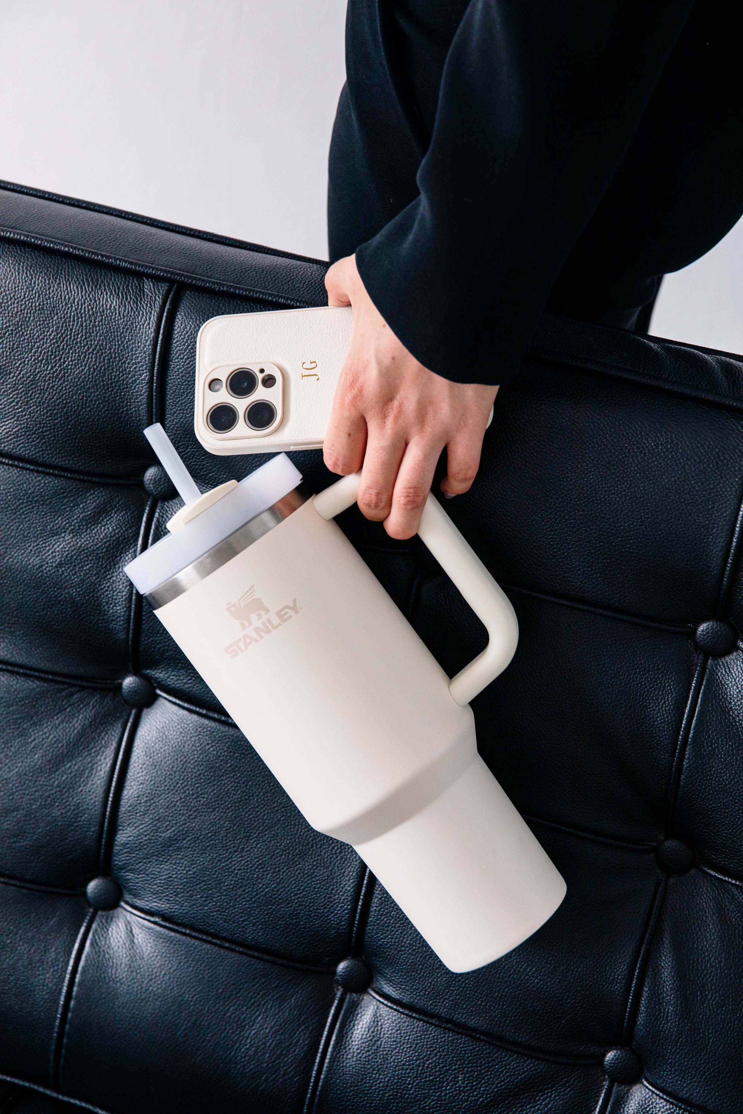 Hand hält weiße Stanley-Thermosflasche mit Strohhalm, daneben liegt ein weißes iPhone mit Monogramm auf schwarzer Ledersofa.