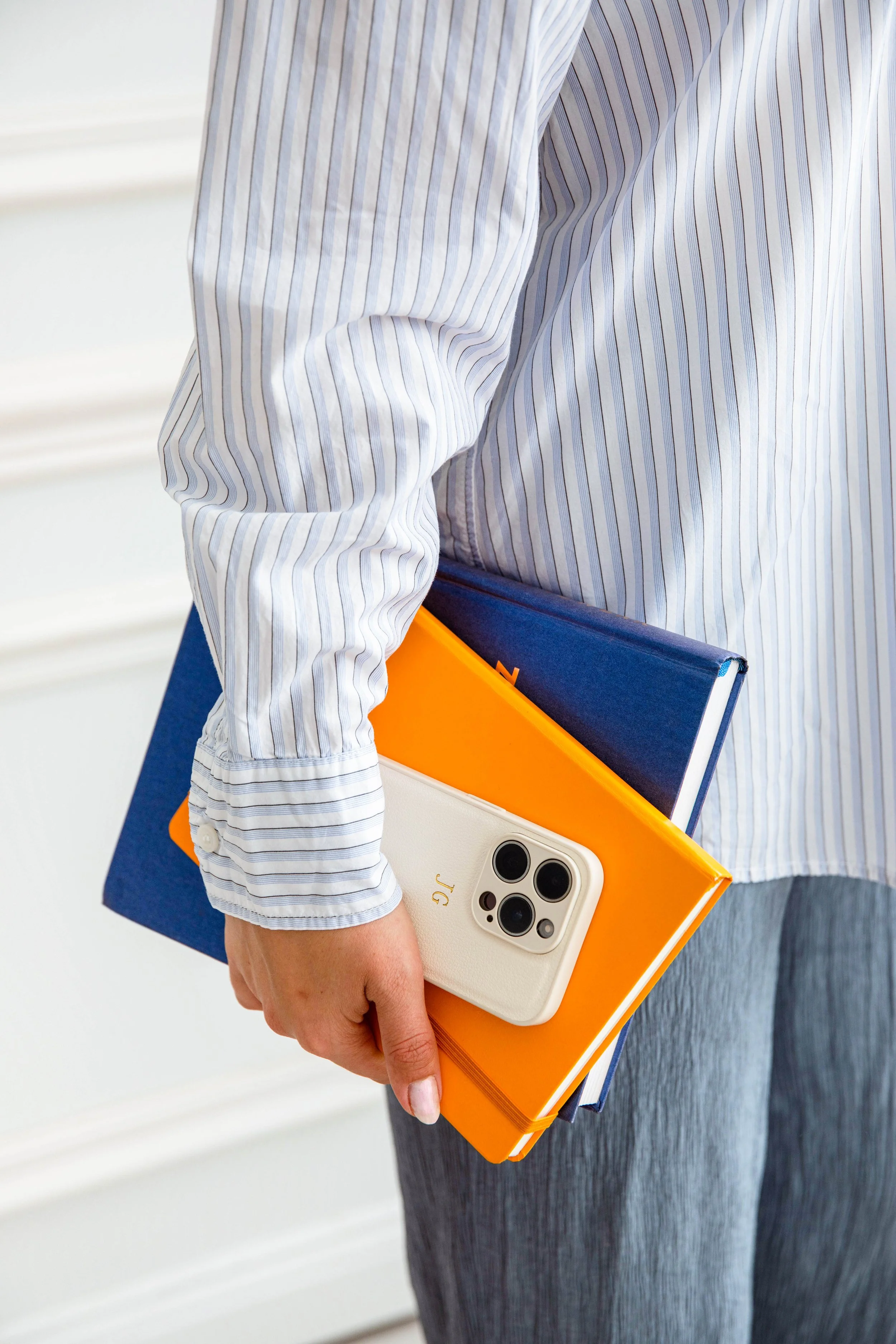 Person hält einen orangefarbenen Notizbuch, ein blaues Buch und ein Smartphone mit weißer Hülle und drei Kameras. Person trägt ein gestreiftes Hemd mit langem Ärmel.