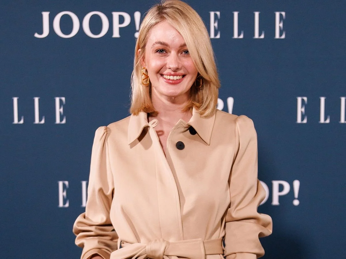 Vanessa Tamkan trägt einen beige Trenchcoat und lächelt vor einem blauen Hintergrund mit weißen Schriftzügen JOOP! und ELLE. 