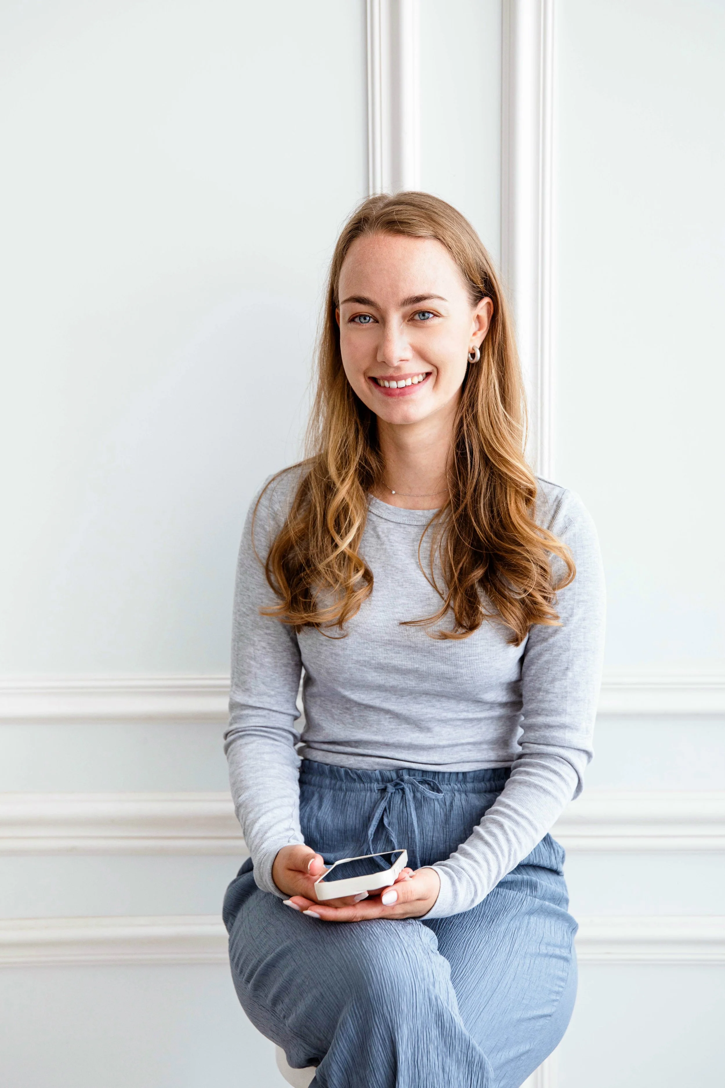 Junge Frau sitzt auf einem Stuhl, hält ein Smartphone in den Händen und lächelt.