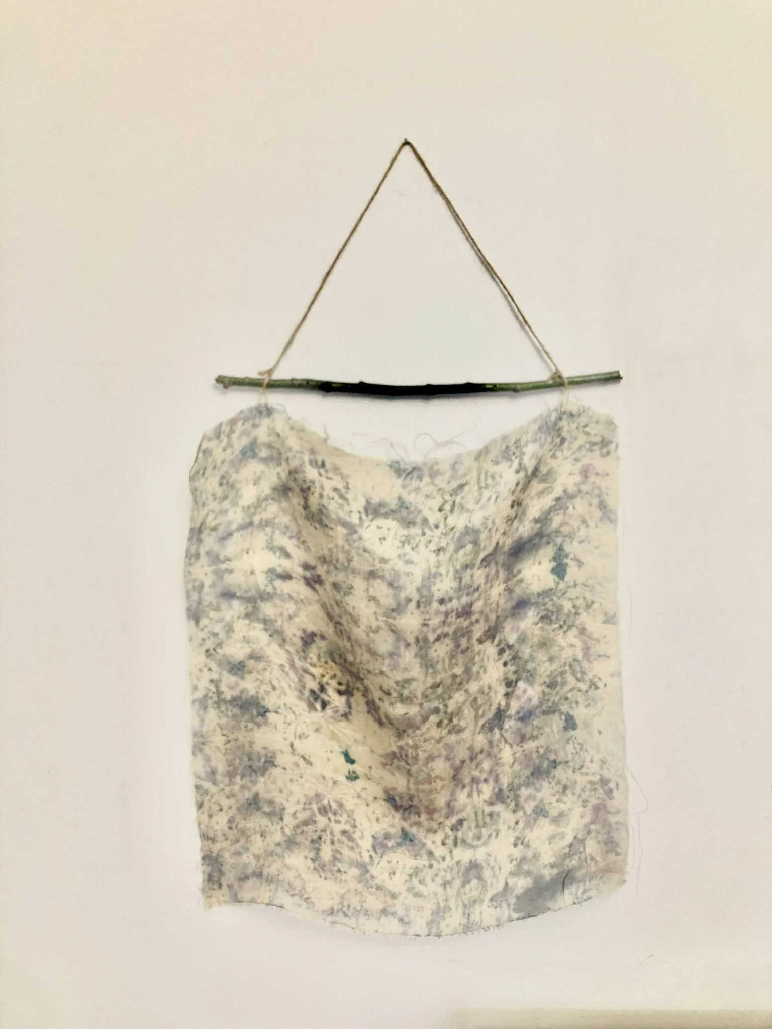 A textured cloth or fabric hanging on a piece of string attached to a stick, hung against a plain white wall.