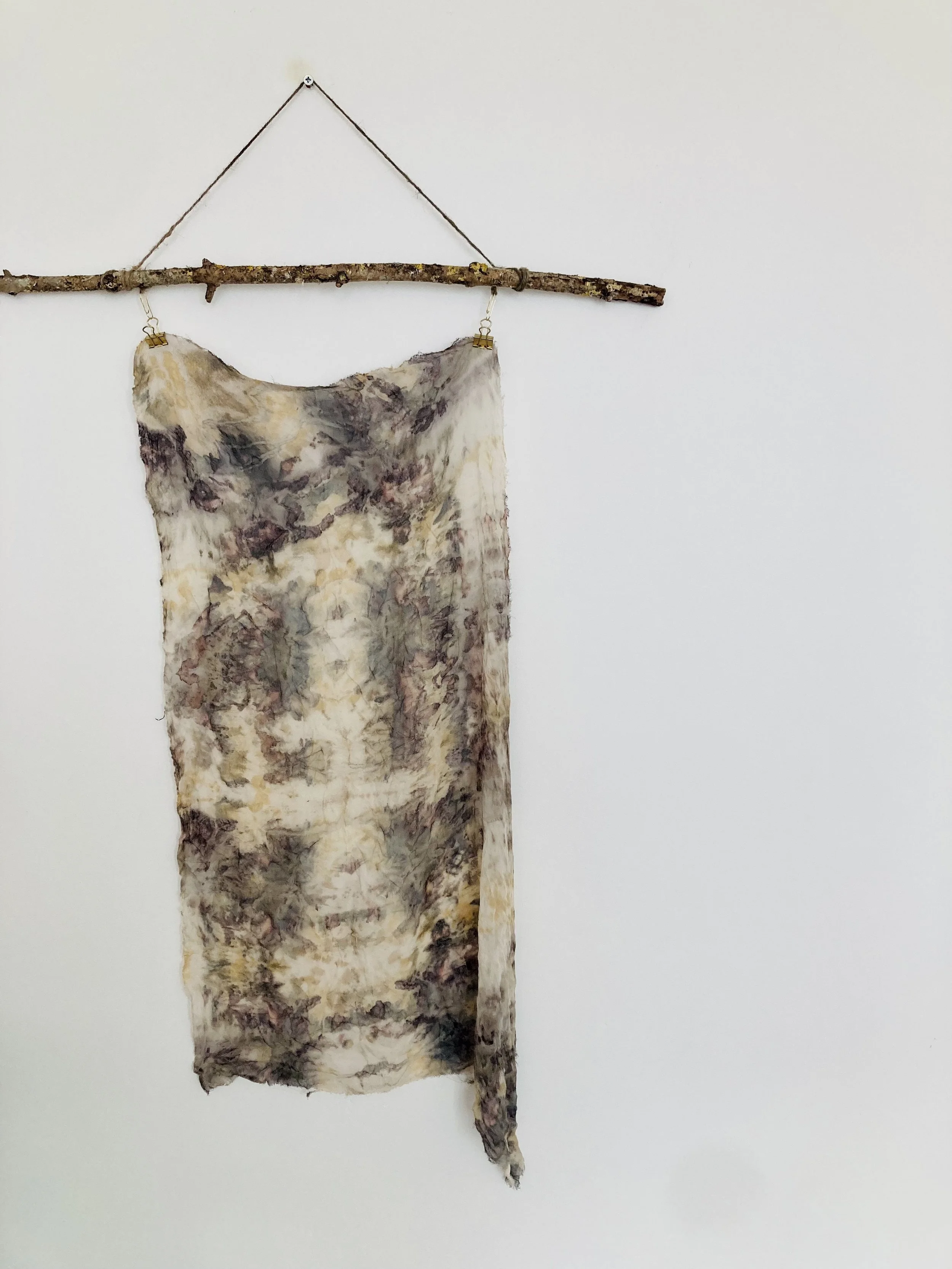 A piece of abstract tie-dye fabric hanging from a wooden stick, suspended by two clips, against a plain white wall.