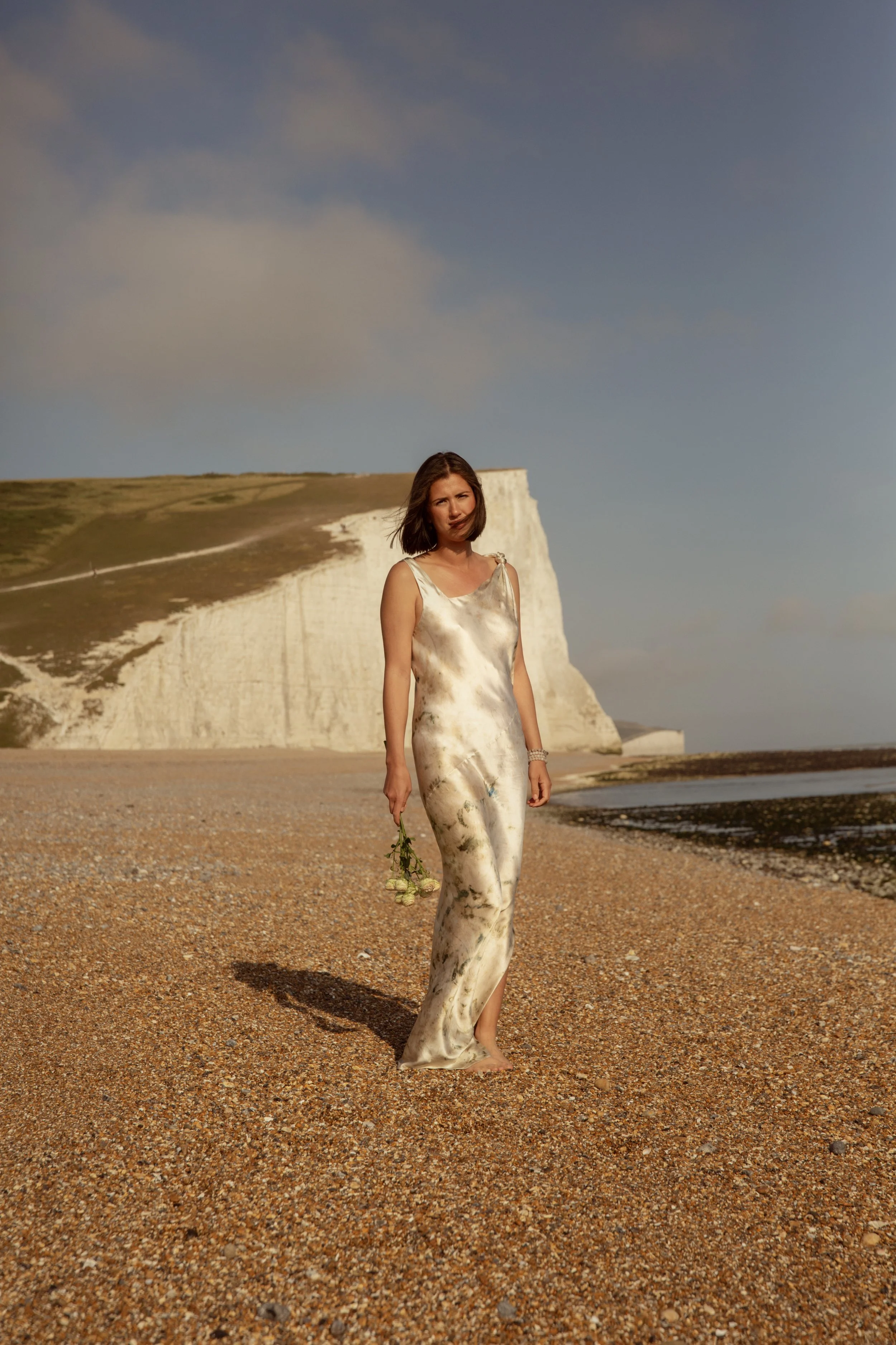 A woman in a long, satin floral dress walking barefoot on a pebble beach, holding a small bouquet of flowers. White chalk cliffs and a partly cloudy sky are in the background.