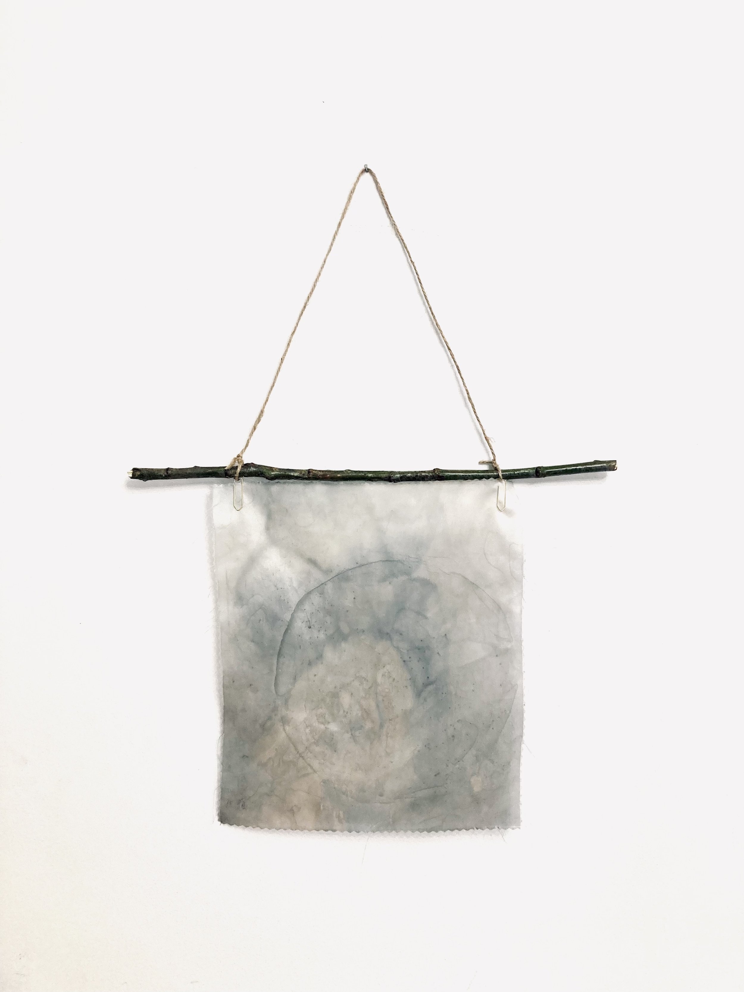 A watercolor artwork featuring a circular design on paper, hanging from a horizontal twig supported by a string attached to a wall.