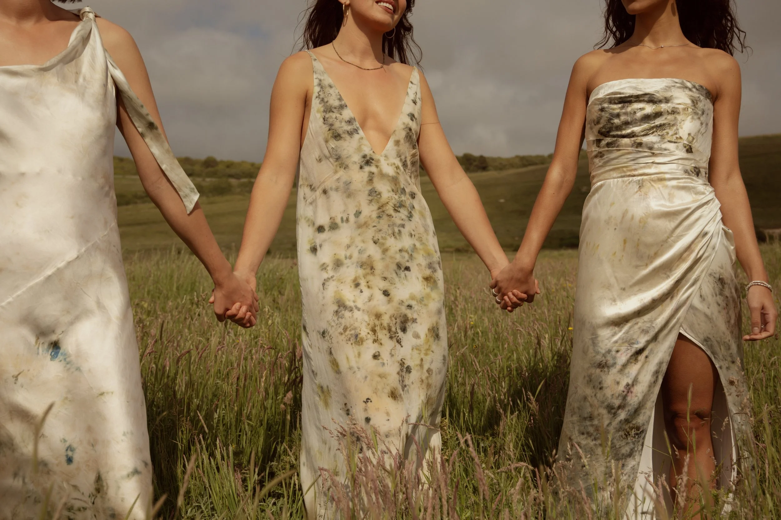 Three women holding hands in a field, wearing tie-dye dresses, with a landscape of rolling hills and a cloudy sky in the background.