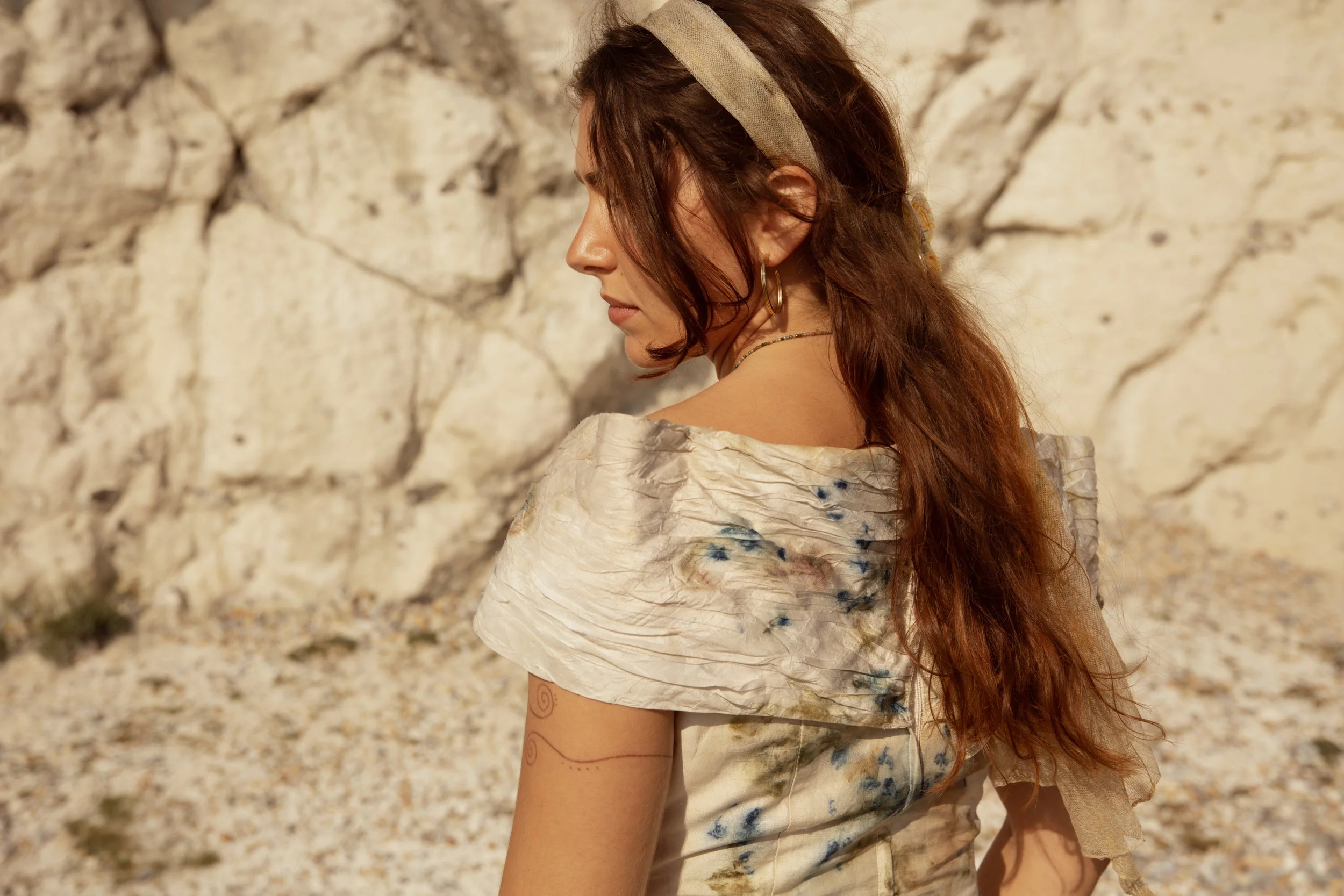 A woman with long red hair, wearing a beige headband and a light-colored off-shoulder dress, standing in front of a rocky background.