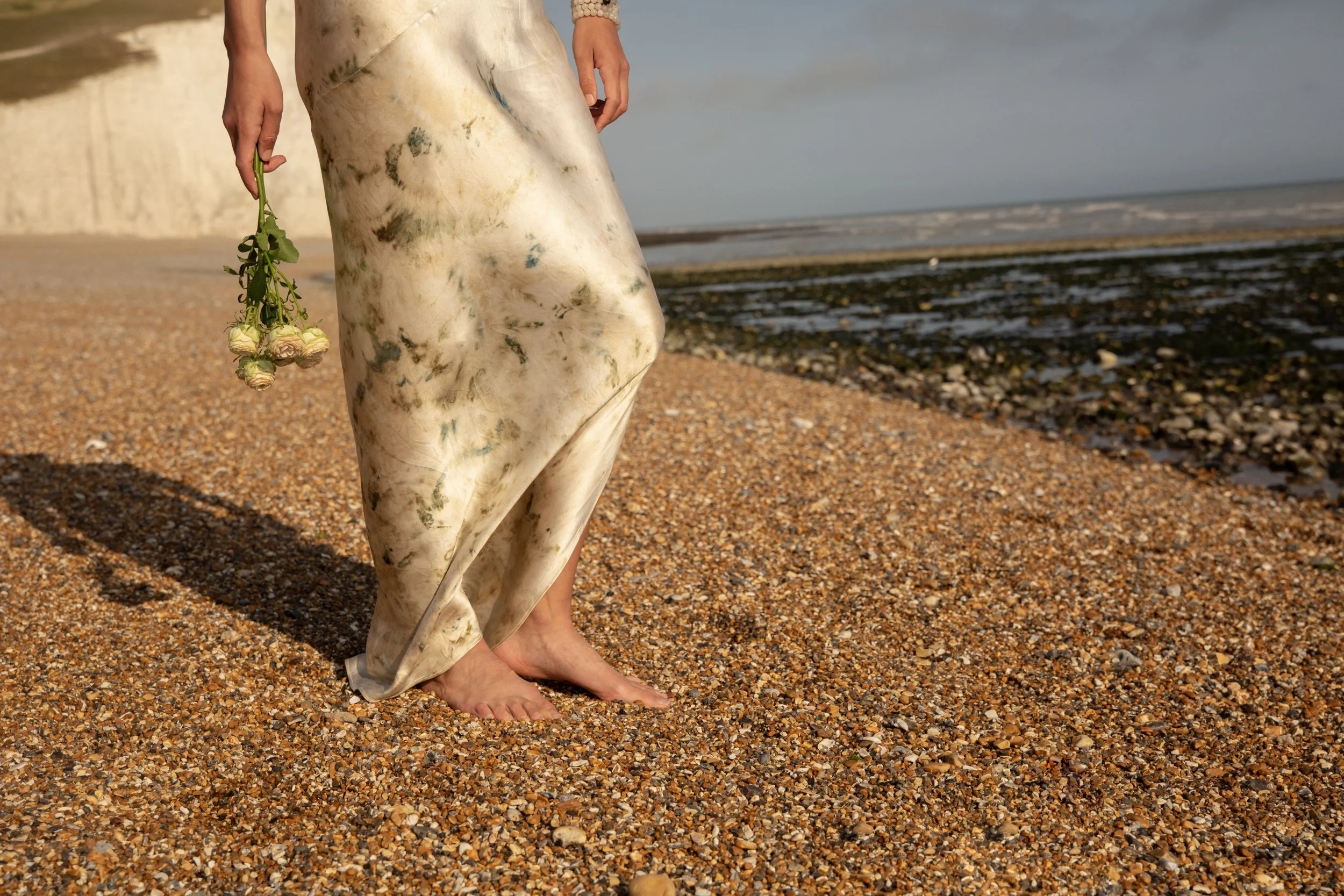 Person standing barefoot on a pebble beach, holding a small bouquet of white roses, with the ocean in the background.