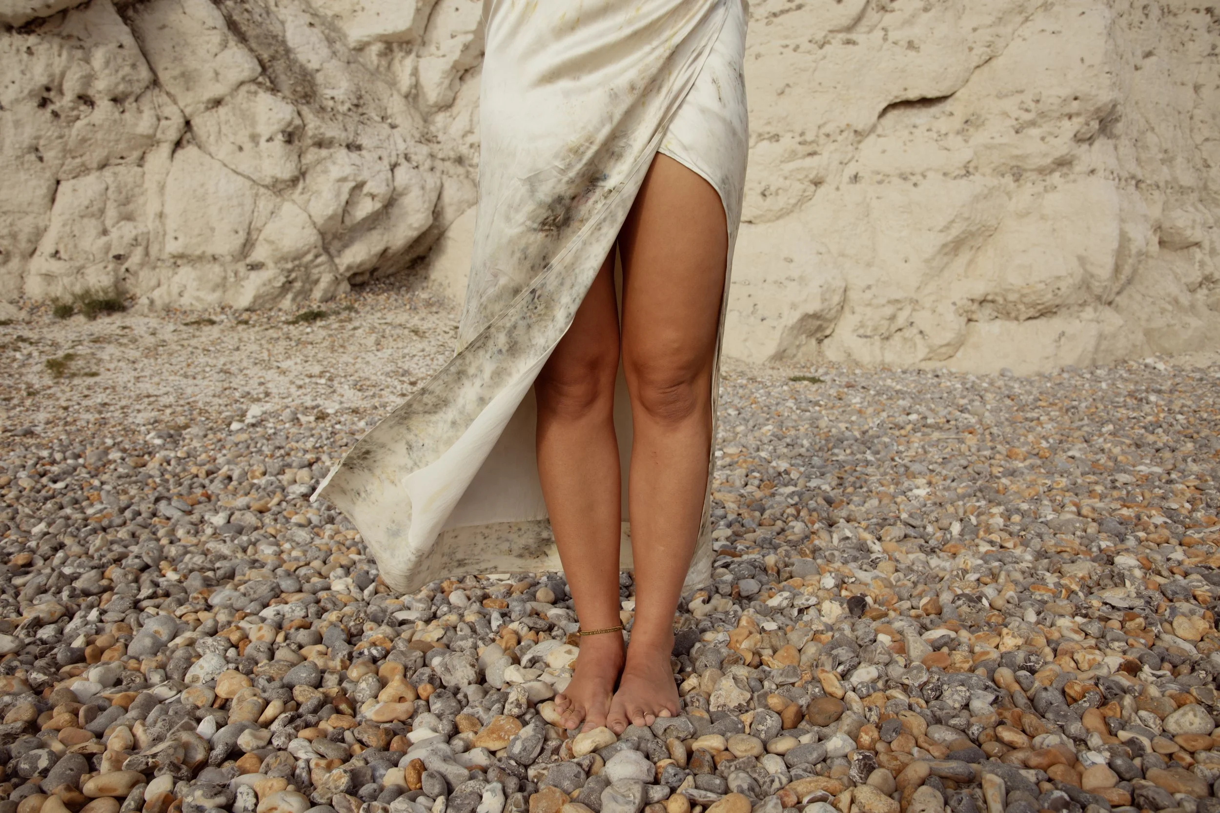 Close-up of a woman's legs standing barefoot on a rocky beach, with a flowing, dirty-white dress covering part of her body and a rocky cliff in the background.
