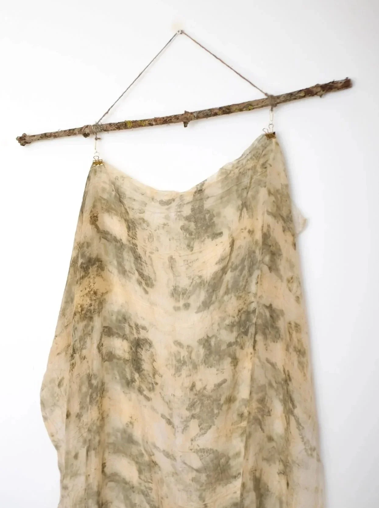 A rustic wall hanging made from a horizontal tree branch with a piece of fabric draped from metal hooks on either end, hanging against a white wall.