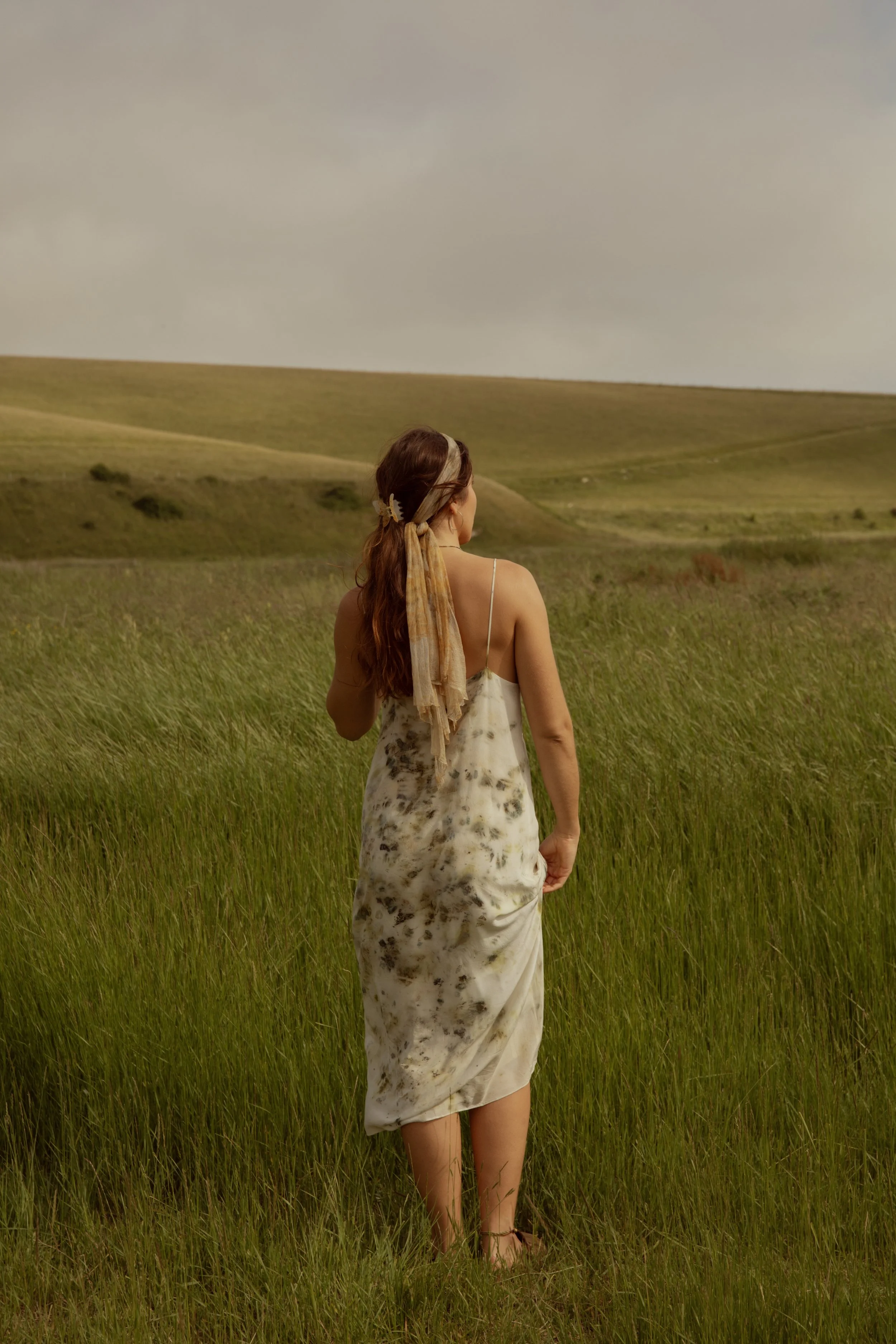 A woman in a white dress with dark stains stands in a grassy field, facing away, with rolling hills in the background under a cloudy sky.