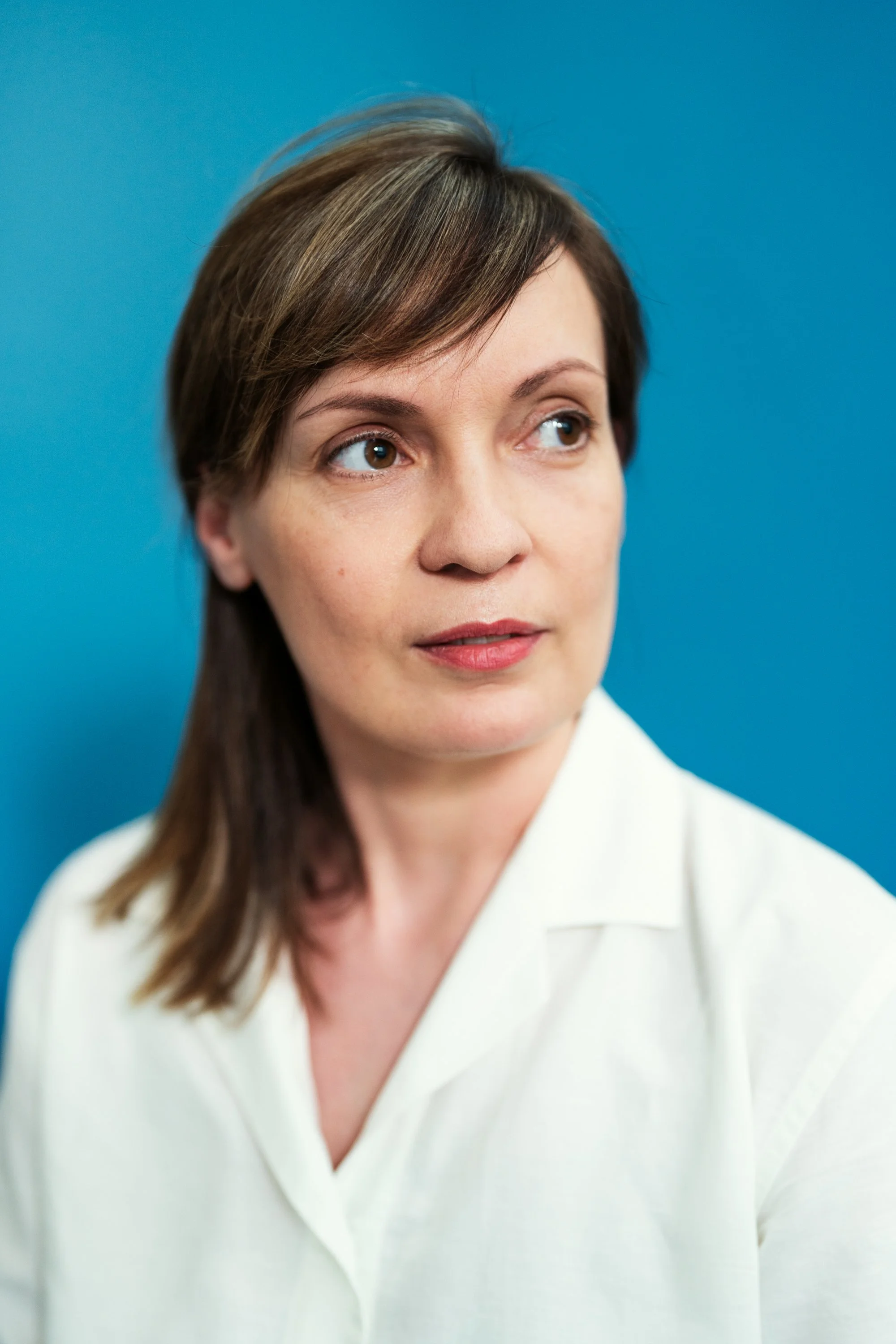 Porträt einer Frau mit braunen, schulterlangen Haaren vor einem blauen Hintergrund, trägt eine weiße Bluse.