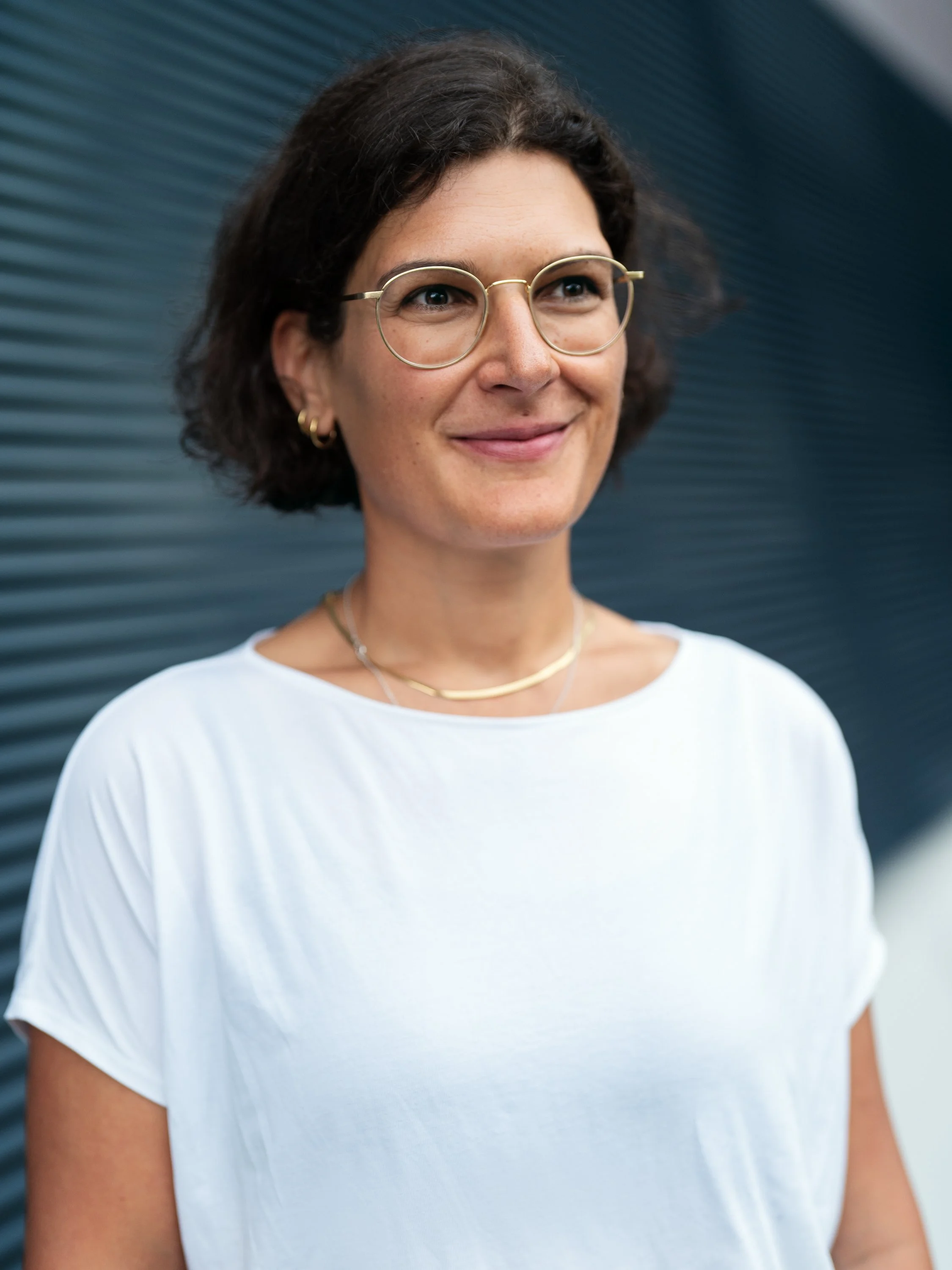 Portrait einer Frau mit dunklen, lockigen Haaren, Brille und weißen T-Shirt vor einer dunklen, modernen Wand.