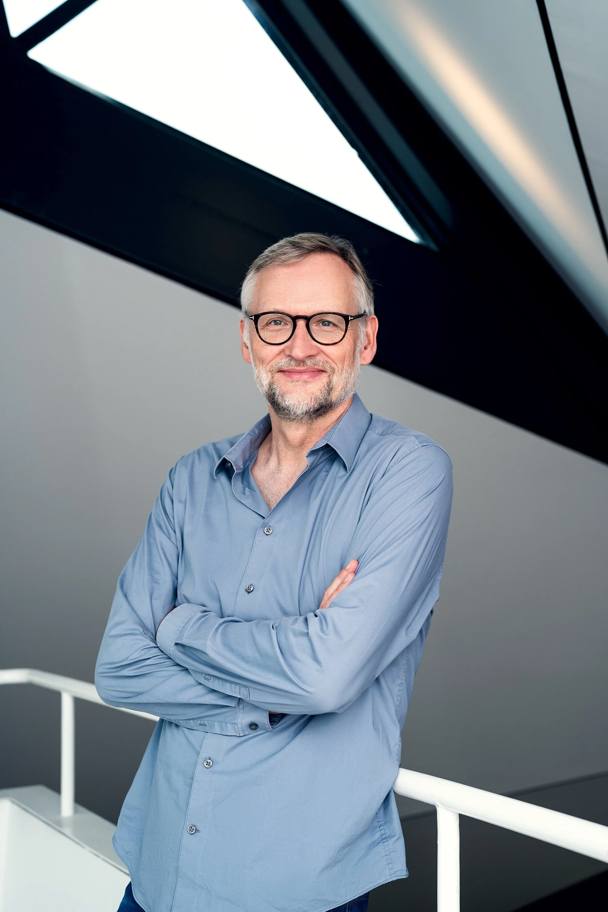Ein Mann mit Brille und grauem Bart steht mit verschränkten Armen vor einer modernen Architektur aus Glas und Stahl.