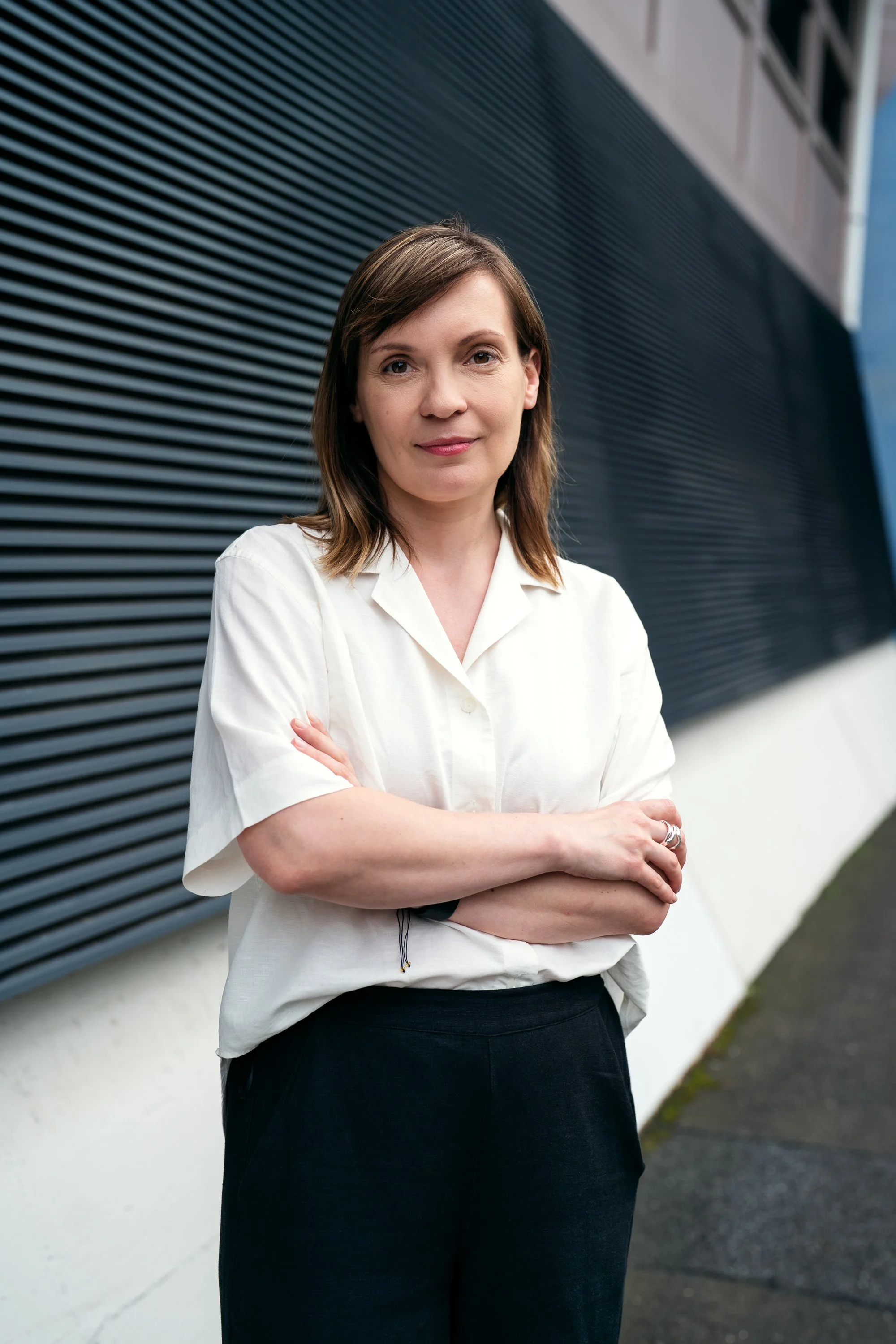 Eine Frau mit braunen Haaren steht vor einem modernen Gebäude mit schwarzen Lamellen an der Fassade, trägt einen weißen Hemd und schwarze Hose, die Arme verschränkt.