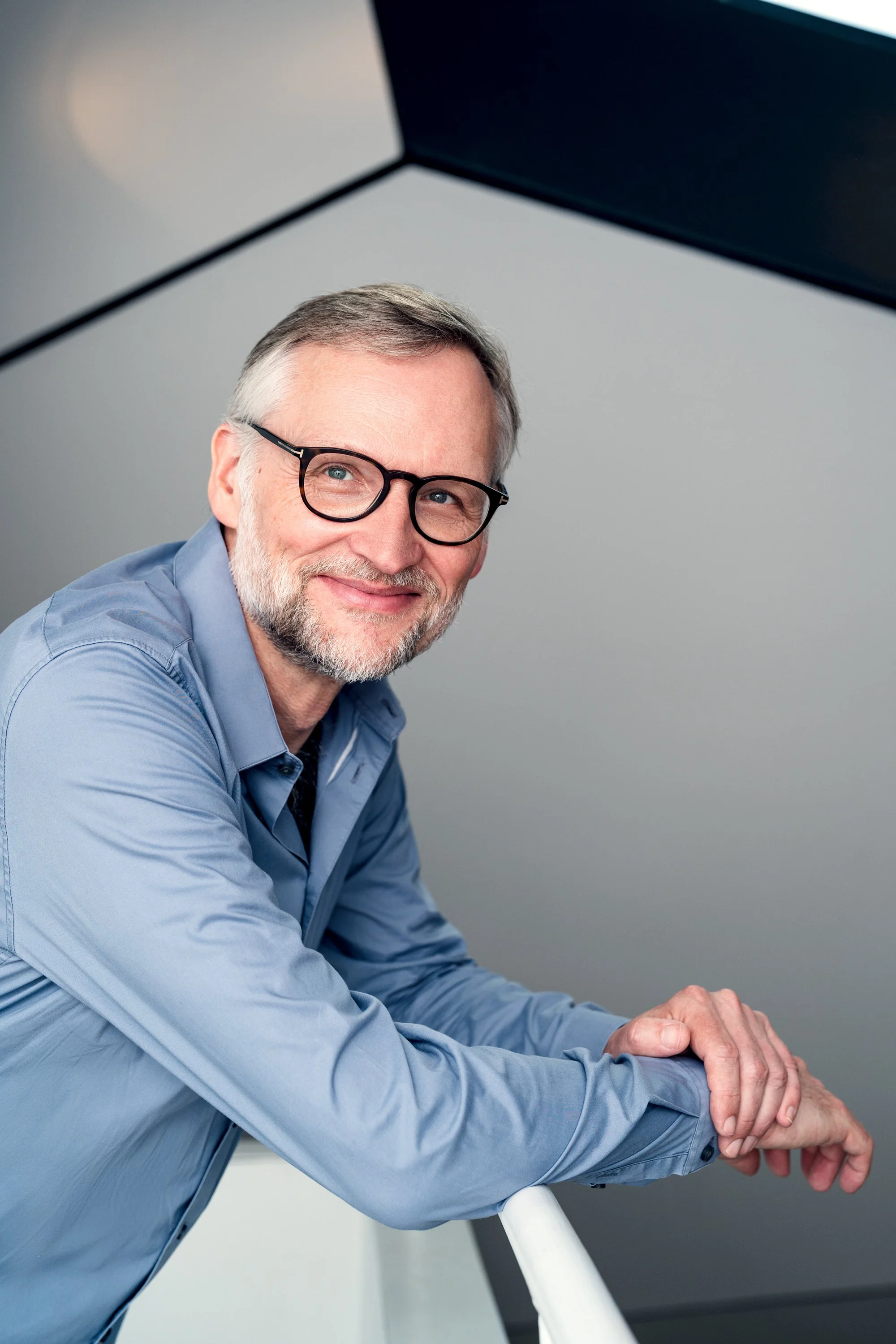 Ein lächelnder Mann mit Brille in einem blauen Hemd sitzt an einem Geländer.