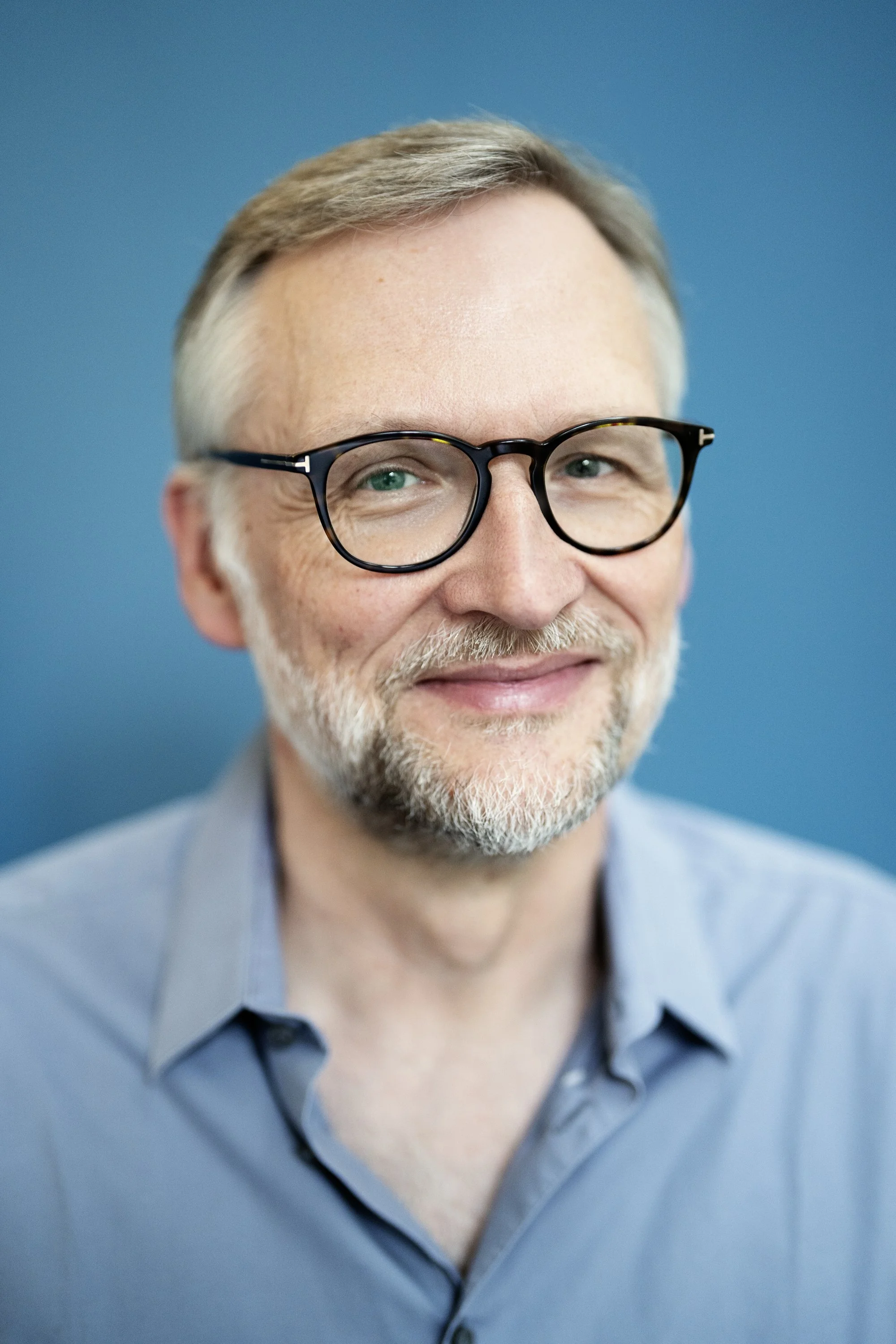 Nahaufnahme eines lächelnden Mannes mit Brille vor blauem Hintergrund.