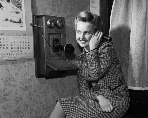 A woman smiling while talking on a vintage wall-mounted telephone in a room with patterned wallpaper and a calendar.