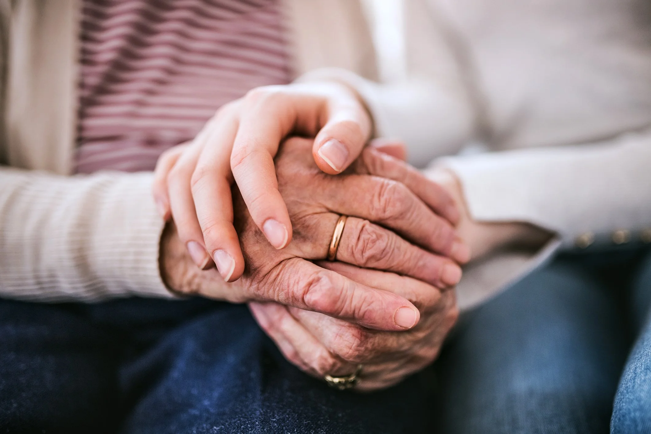 Close-up of two hands, one elderly and one younger, gently holding each other, with the elderly hand wearing a gold wedding band.